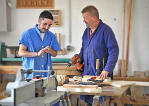 2 Männer in einer Werkstatt. Ein Mann ist älter als der andere. der Junge mann macht etwas mit Holz und der ältere schaut zu während er eine Bohrmaschine in der Hand hält. Überall ist viel Holz und Werkzeug