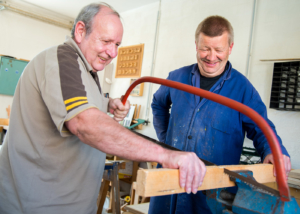 Mann sägt in Werkstatt ein Holzbalken durch. Ein Handwerker schaut begeistert zu.
