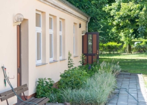 Ein Foto von einer gebäudefassade in einer schrägen Ansicht. Die Fassade ist beige und hat 4 Fenster und 2 braune Türen. Eine Tür ist geöffnet. Vor dem Gebäude ist eine Sitzbank und ein schönes Pflanzenbeet. Im Hintergrund kann man viele Bäume und Büsche erkennen