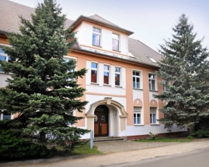 Das Haus am Berghang. Ein schönes historisches Gebäude mit viel Charakter in Lehmfarben und vielen Strebenfenstern. Große Nadelbäume befinden sich deneben.
